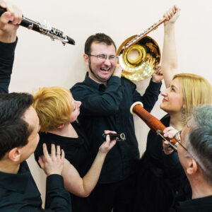 novosadski duvački kvintet, na slici nekoliko muzičara drži svoje instrumente prema jednom koji stoji u centru i jako se smeje svojim kolegama