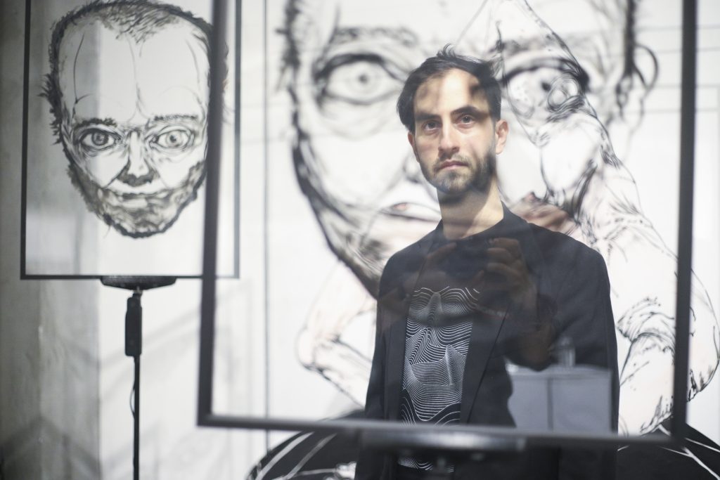 umetnik stoji ispred projekcije svojih crteža koji se oslikavaju po njegovom licu i iza njega na belom platnu